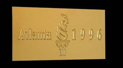 The metal nameplate is a gold-colored plaque, prominently displaying the text "Atlanta 1996." Centered between the city name "Atlanta" and the year "1996," there is a stylized Olympic torch with a vibrant flame, symbolizing the spirit of the Games. Unlike typical company-branded nameplates, this one commemorates the 1996 Summer Olympics held in Atlanta, emphasizing its historical significance rather than associating it with a specific business or manufacturer. The design is both elegant and prestigious, befitting an event of global importance.