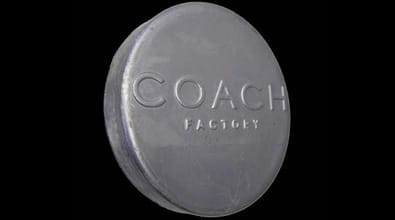 The metal nameplate is circular and made of metallic material, featuring the company name "COACH FACTORY" embossed in raised lettering. The polished finish of the plate catches the light, creating a subtle shine that contrasts elegantly against its black background. The prominent "COACH FACTORY" inscription serves as a clear identifier, showcasing the brand’s signature style through its refined craftsmanship and minimalistic design.