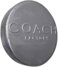 The metal nameplate is circular in shape and made of silver-toned metal. The words "COACH FACTORY" are prominently embossed on the surface, giving them a slightly raised texture that catches the light. The overall finish of the nameplate appears smooth and polished, adding a touch of shine against a contrasting black background. This style of nameplate is typically used by the COACH brand to indicate authenticity or branding on their factory line products.