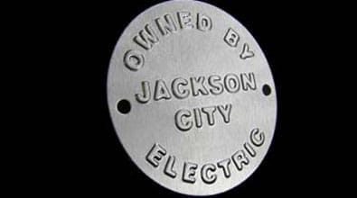 This is a round, silver-colored metal nameplate designed for identification purposes. At its center, raised (embossed) lettering spells out “OWNED BY JACKSON CITY ELECTRIC” in capital letters, creating a bold and prominent display. The inscription stands out against a contrasting black background, making the text highly legible. On either side of the central area are two small holes—most likely for mounting the nameplate onto equipment or property owned by Jackson City Electric. The overall look suggests an industrial or utility use, emphasizing both durability and clear ownership by the company “Jackson City Electric.”.