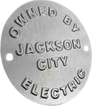This is a round metal nameplate featuring two small mounting holes, likely for screws or rivets, positioned opposite each other near the edge. The face of the nameplate is engraved in bold uppercase letters with the text: “OWNED BY JACKSON CITY ELECTRIC.” The engraving clearly displays the company’s name—Jackson City Electric—indicating ownership or identification for equipment or property. The metallic finish stands out against a plain black background, accentuating the crisp lettering and simple industrial design typical of utility or municipal asset tags.