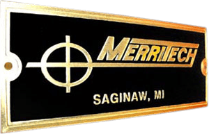 This is a rectangular metal nameplate with a sleek black background bordered by a gold outline. Prominently displayed in gold lettering is the company name “MERRITECH,” which is positioned next to a distinctive crosshair symbol, emphasizing precision or targeting. Beneath the main text, “SAGINAW, MI” indicates the company’s location. The design includes two screw holes on either side for easy attachment, making it both decorative and functional as an industrial or equipment identification plate. The combination of black and gold gives the nameplate a professional and high-quality appearance appropriate for business branding or machinery marking.