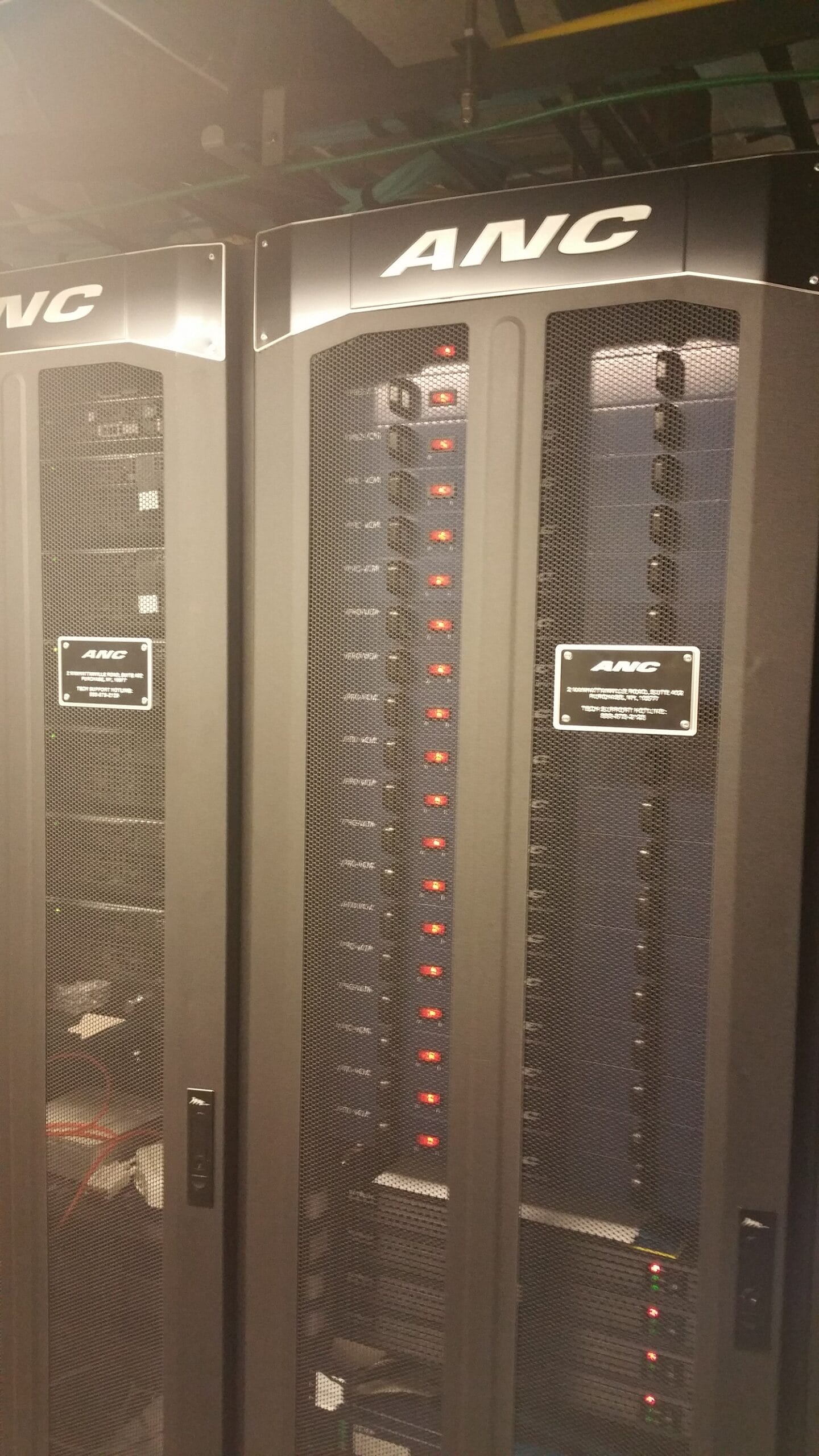 The metal nameplate on the server rack prominently displays the company name "ANC" in bold, likely engraved or embossed letters. It is affixed to the front of the rack, standing out clearly even in the dimly lit room. Positioned against a backdrop of glowing red indicator lights visible through a mesh door, this sturdy metallic plaque serves both as an identifier and a mark of ownership or manufacture—signaling that ANC is either the manufacturer or operator of this equipment. The overall appearance conveys professionalism and high-tech reliability.