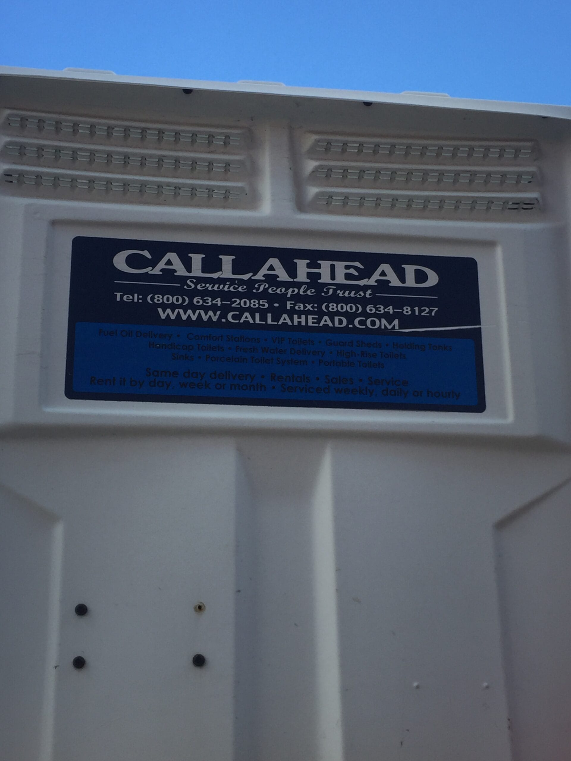 The metal nameplate on the white portable toilet is prominently blue in color and features the company name "CALLAHEAD" in bold white lettering. Beneath the company name, additional contact information, including a phone number and website address, is clearly displayed in white text. The blue background of the nameplate also lists a range of services offered by CALLAHEAD, making it easy to identify the provider and access their services. The vibrant contrast between the blue nameplate and its white text ensures high visibility. Above the unit, an open sky can be seen.