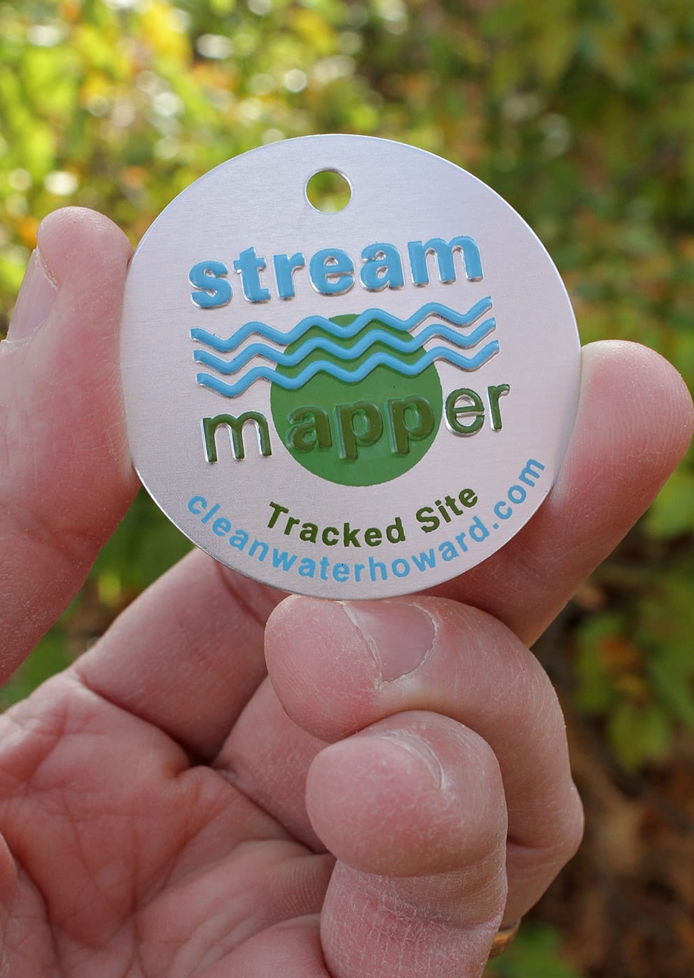 A hand holds a round metal nameplate tag prominently featuring the company name "Clean Water Howard," as indicated by the etched website cleanwaterhoward.com. The tag’s design includes the title "stream mapper" at the center, with wavy blue lines and a green circle evoking a natural water theme. Additional text reads "Tracked Site," all etched into the metal. The background is softly blurred greenery, highlighting that this durable, engraved tag is likely used outdoors to signify sites tracked or protected by Clean Water Howard’s environmental initiatives.