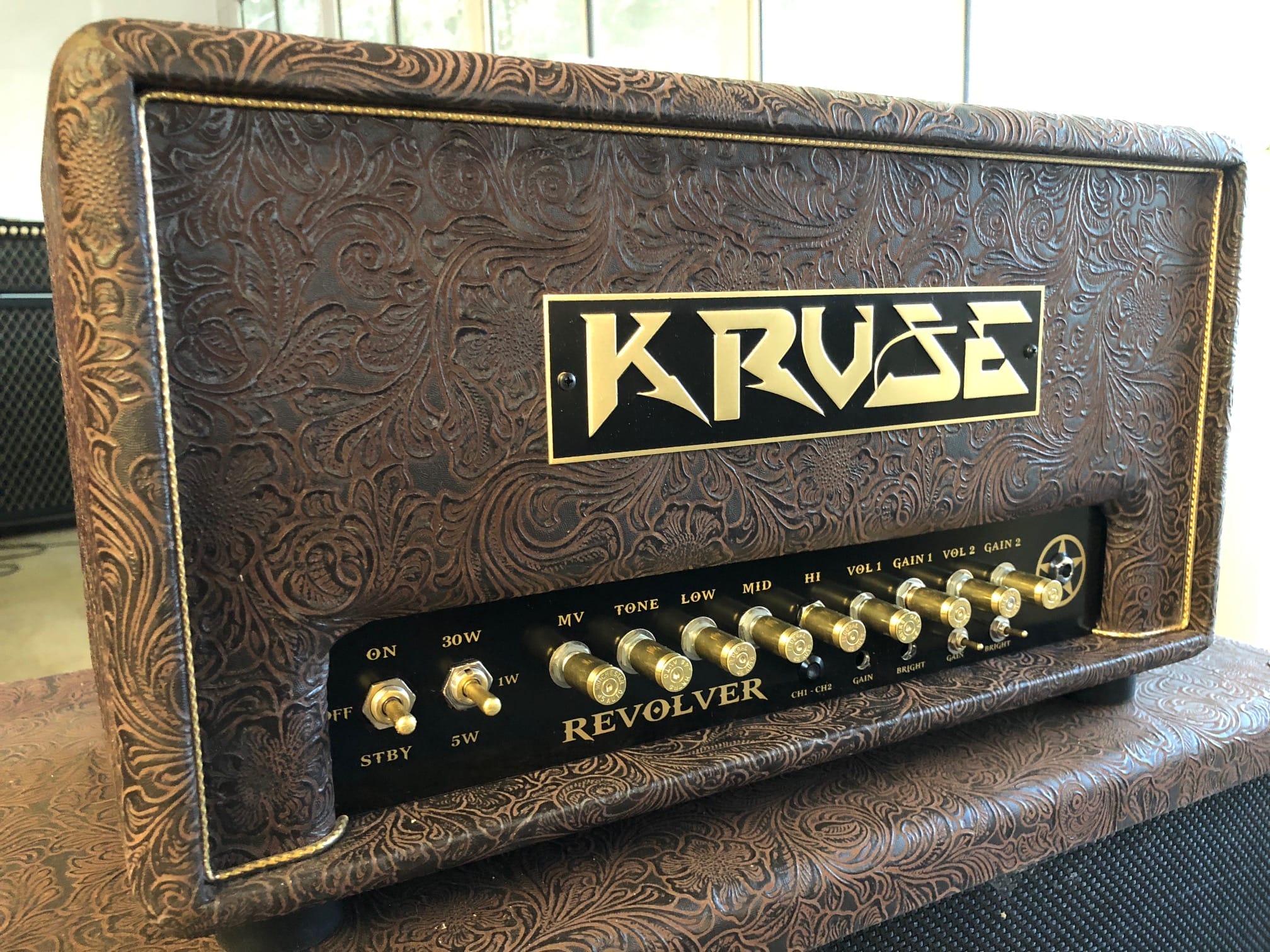 The metal nameplate on the Kruse Revolver guitar amplifier head prominently features the company’s name “Kruse.” The nameplate is likely rectangular and affixed to the front panel, standing out against the amplifier’s ornate, textured brown casing. Crafted from metal with a polished finish, it serves as a bold identifier for the brand. The lettering "Kruse" is centrally displayed—possibly in a stylish or classic font—ensuring instant recognition. This branded plate is positioned above or near the gold control knobs and switches, providing an elegant and professional accent to the overall vintage aesthetic of the amp head.