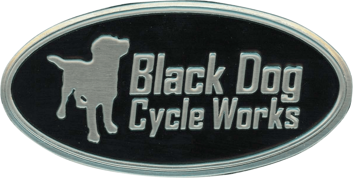 This is an oval-shaped metal nameplate featuring the bold, embossed company name "Black Dog Cycle Works." The design includes a prominent silhouette of a dog, emphasizing the brand's identity. The combination of sturdy metal construction, raised lettering, and distinctive dog graphic gives the nameplate an industrial yet personalized look that's perfect for branding motorcycles or related equipment.
