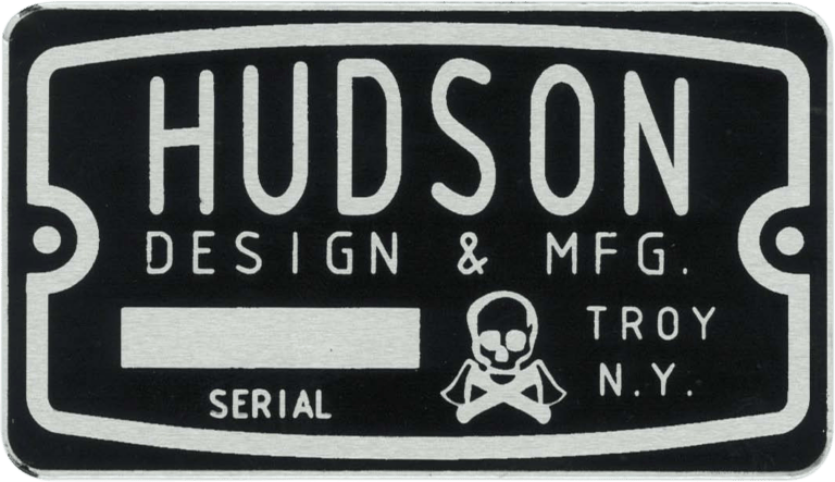 The metal nameplate is rectangular and made from durable black and silver metal. It features the company name "HUDSON DESIGN & MFG." prominently at the top, followed by the location "TROY N.Y." Beneath this, the word "SERIAL" is printed, next to a blank space intended for a serial number. At the bottom of the nameplate, there is a skull and crossbones symbol, suggesting caution or danger related to the equipment or product it identifies. The overall appearance conveys an industrial or manufacturing context and implies that HUDSON DESIGN & MFG. is responsible for its production in Troy, New York.